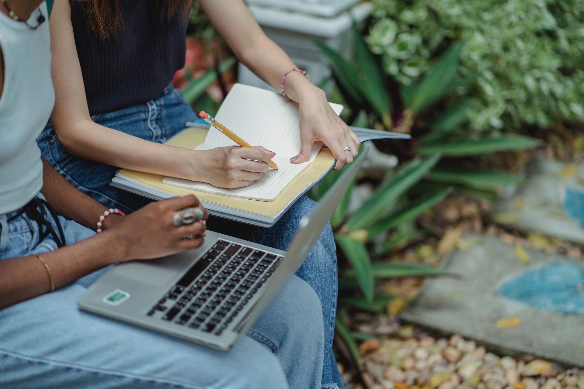 Two women seated closely together working outdoors on a laptop and writing in a notebook 