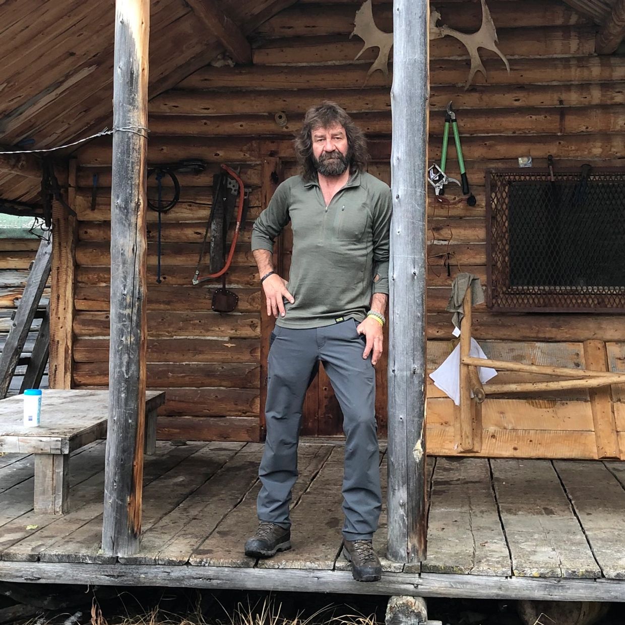 Bearded man standing on a rustic wooden porch of a log cabin.