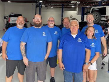 Group of seven smiling mechanics in blue shirts at a garage.
