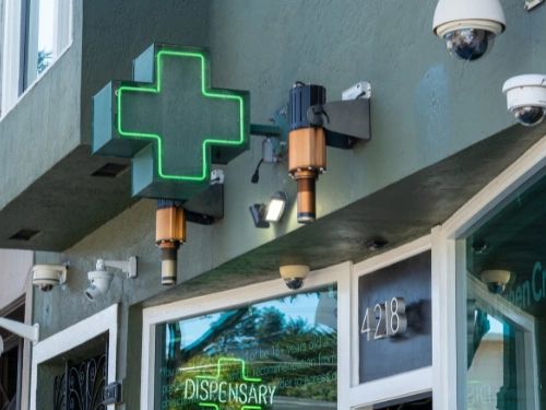 Exterior of a dispensary with a green cross sign and multiple security cameras.