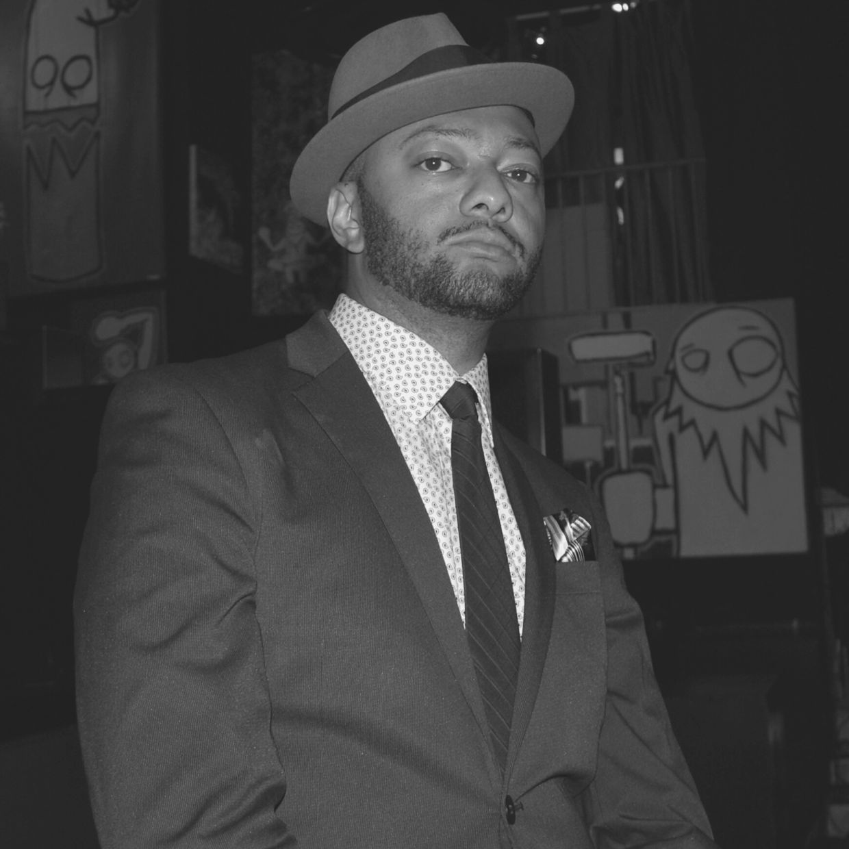 Man in a suit and hat posing confidently indoors with abstract art in the background.