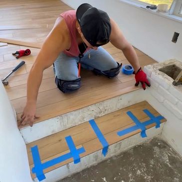 Person installing wooden flooring on a step, using blue painter's tape for alignment.