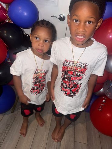 Two boys in matching birthday outfits stand among colorful balloons.