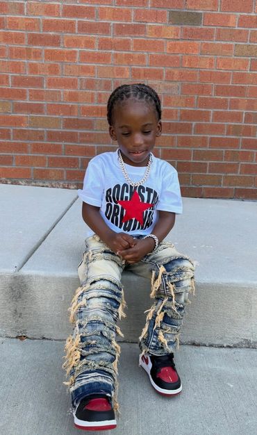 Young boy in stylish ripped jeans and a graphic tee sitting on a concrete curb.