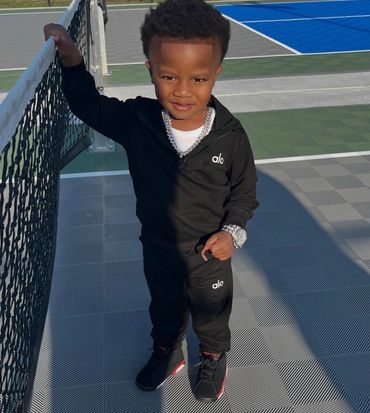 A young boy in black athletic wear stands by a tennis net on a court.