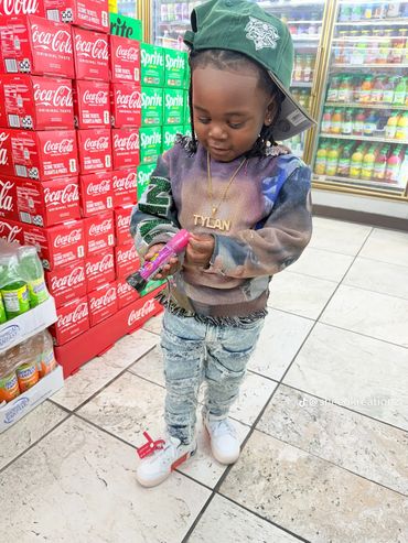 A young child in a colorful hoodie and cap stands in a store aisle with soda boxes.