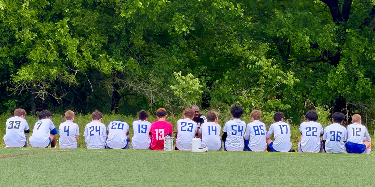 Youth soccer team sitting on grass, backs facing camera, wearing numbered jerseys.