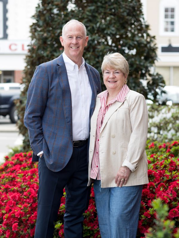 A smiling man and woman stand together in front of bright red flowers outdoors.