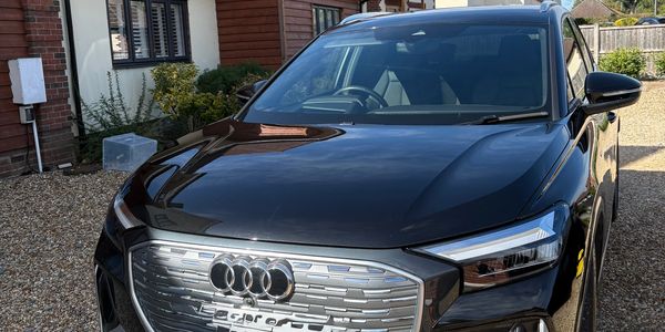 Black Audi e-tron parked on gravel in front of suburban houses under blue sky.