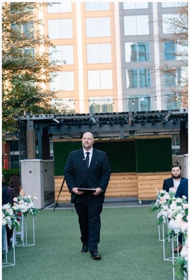 Man in a black suit walking down an outdoor aisle at a formal event.
