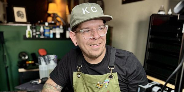 A man wearing a KC cap and green apron smiles in a cozy indoor setting.