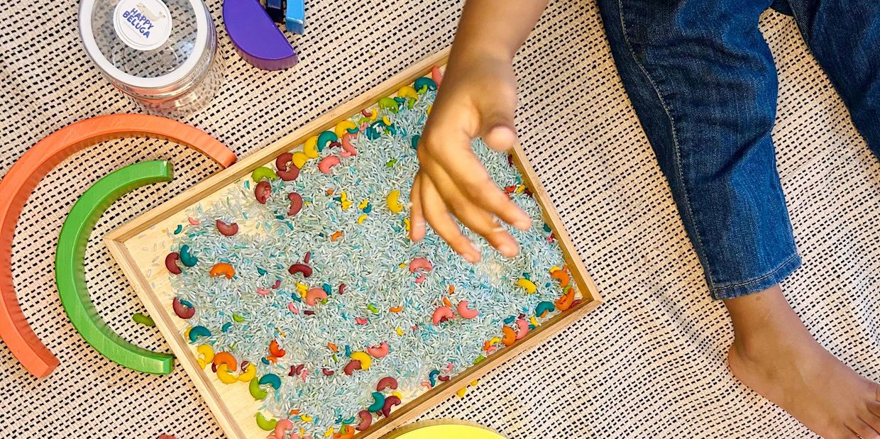 A child playing with colorful sensory toys and a tray of mixed rice and pasta on a textured mat.