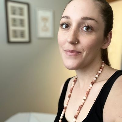 Woman with a nose ring and beaded necklace smiles softly indoors.