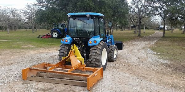 Blue tractor with orange land leveler on a gravel path in a rural setting.