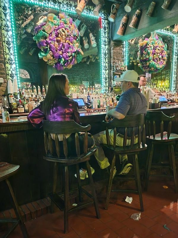 Two people sitting at a bar with colorful decorations and bottles behind.