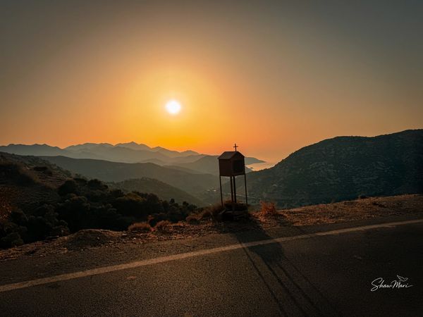 Sunset over mountains with a water tank silhouette by the roadside.
