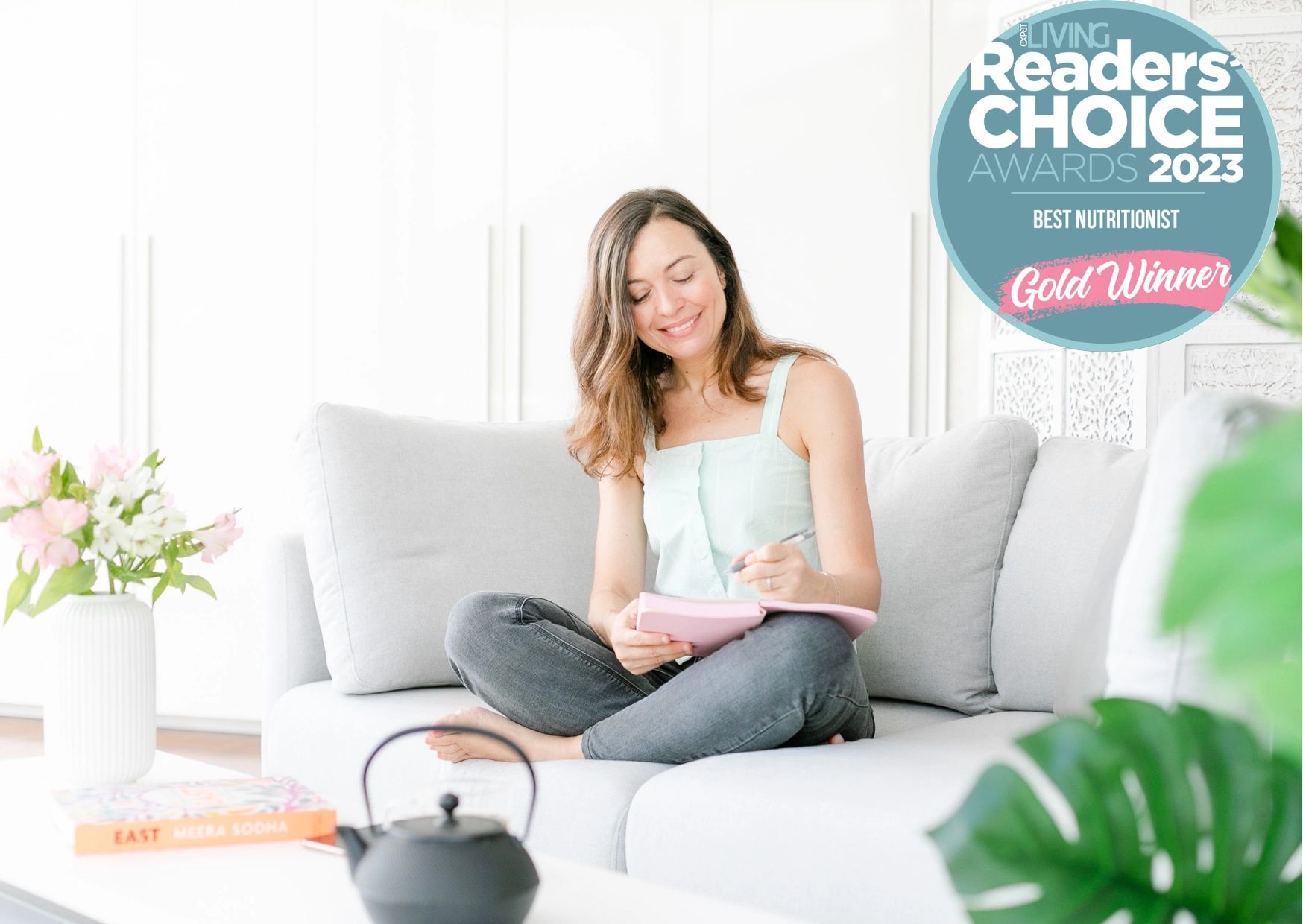 Woman sitting on a couch writing in a notebook, surrounded by a cozy, bright living room.