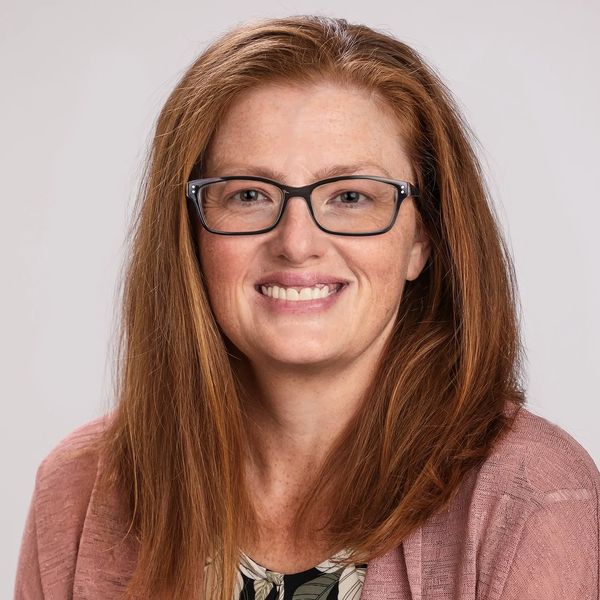 Smiling woman with glasses and reddish hair wearing a floral blouse and pink cardigan.