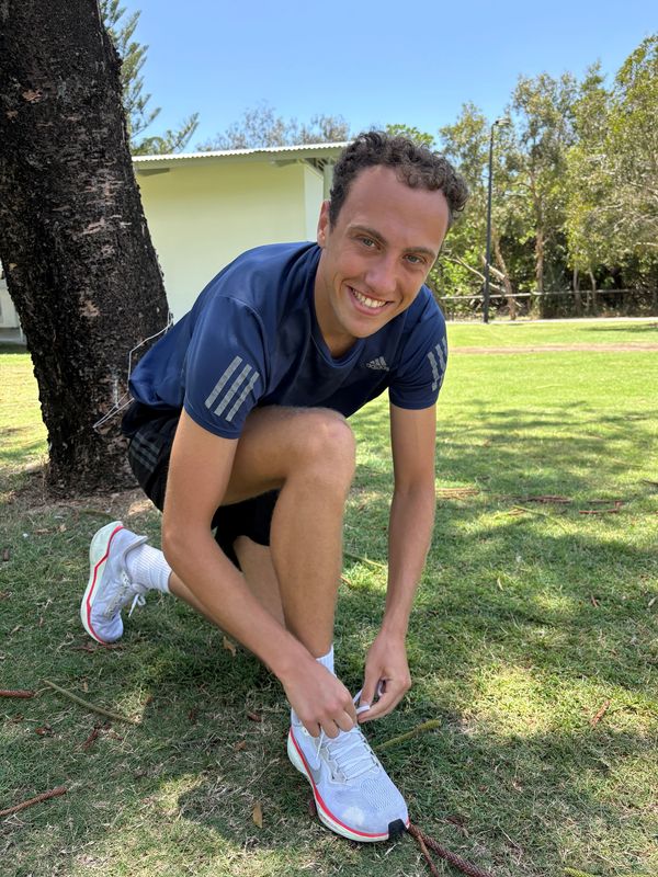A man tying his running shoe on grass near a tree, smiling.