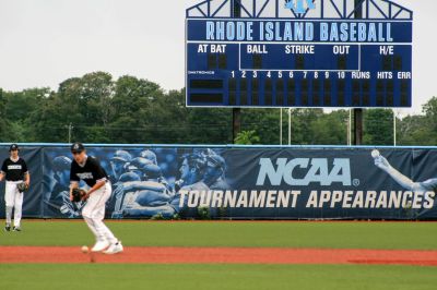 College baseball players on the field during a Rhode Island game with NCAA tournament banner.