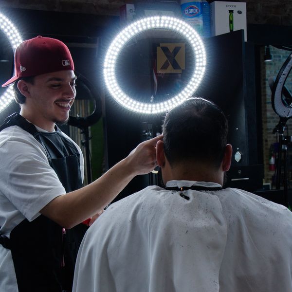 Barber smiling while cutting a client's hair under bright ring light.
