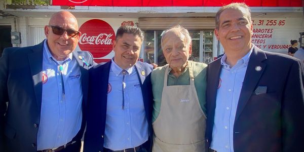 Four men smiling together outside a store with Coca-Cola signage, one wearing an apron labeled Javier.