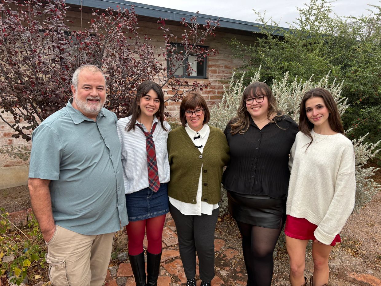 A smiling family of five posing outdoors in front of a brick wall and plants.
