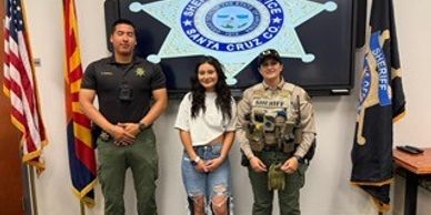 Three law enforcement officers posing in front of a Santa Cruz County Sheriff emblem.