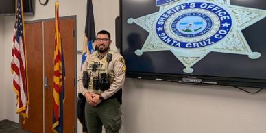 A sheriff officer standing next to flags and a Santa Cruz County badge display.