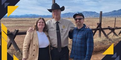 Three people posing outdoors in a rural area with mountains in the background.
