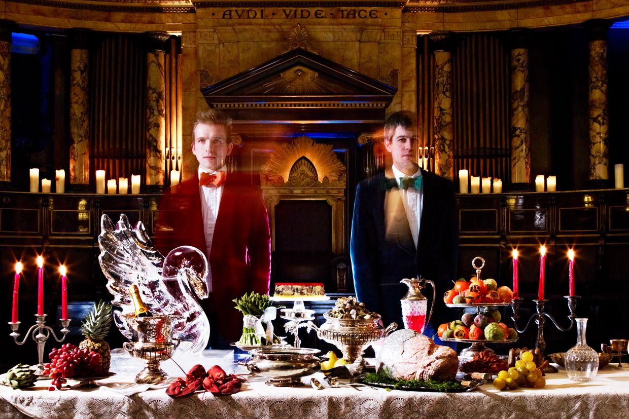 Two men in tuxedos stand behind a lavish banquet table with candles.