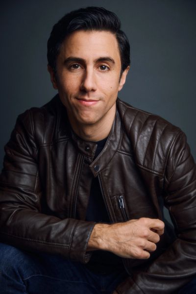 Man in a brown leather jacket posing confidently with a slight smile.