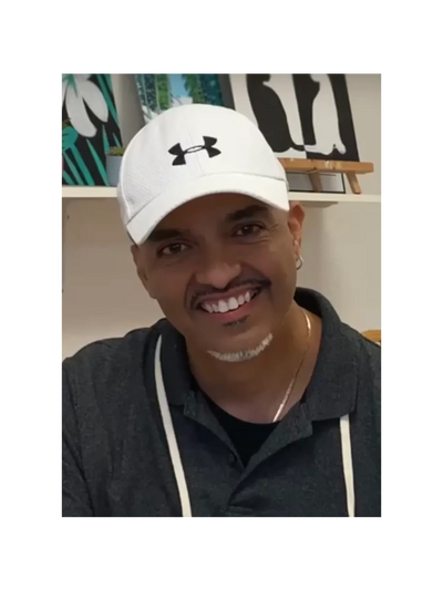 Man smiling, wearing a white Under Armour cap and dark shirt indoors.