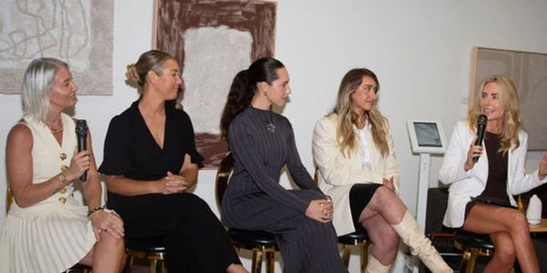 Five women engaged in a panel discussion indoors, two holding microphones.