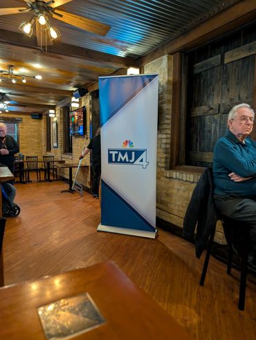 Interior of a rustic venue with a TMJ4 banner and people inside.