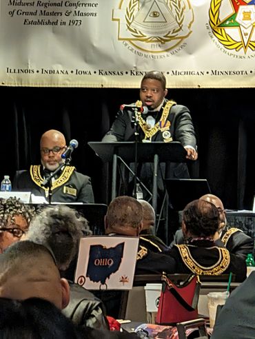 A speaker addressing a Masonic conference with members seated and listening.