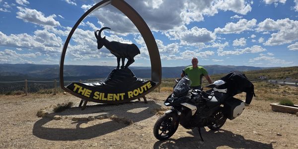 Motorcyclist posing by a sign for The Silent Route under a bright sky.