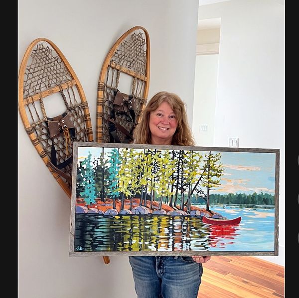 A smiling woman holding a colorful landscape painting in front of vintage snowshoes.