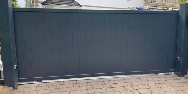 Modern dark gray sliding gate in front of a white house with stone driveway.