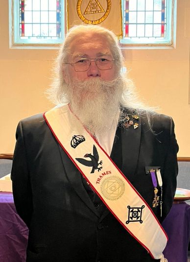 Elderly man with long white beard and ceremonial sash indoors.
