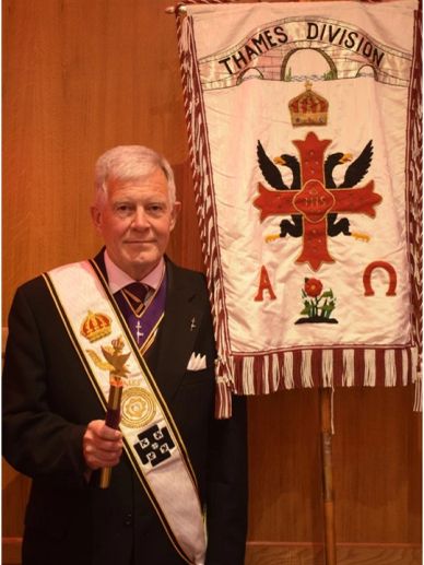 Elderly man in ceremonial attire holding a staff next to a banner.