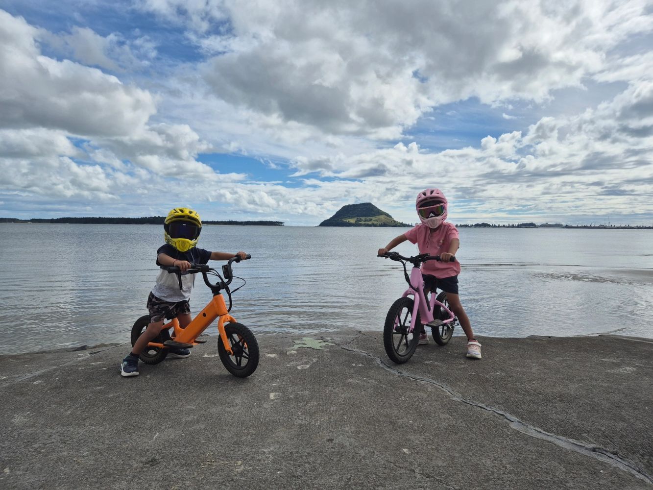 Two kids with helmets on bikes by the water under a cloudy sky.