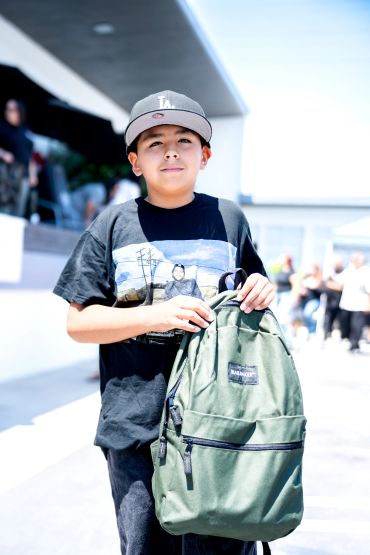 Boy in black shirt and cap holding a green backpack.