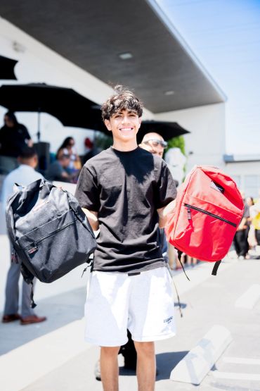 Young man smiling holding two backpacks outdoors.