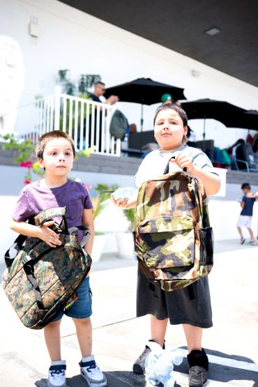 Two boys holding camouflage backpacks outdoors.