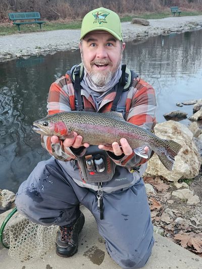 Man proudly holds a large rainbow trout near a riverbank.