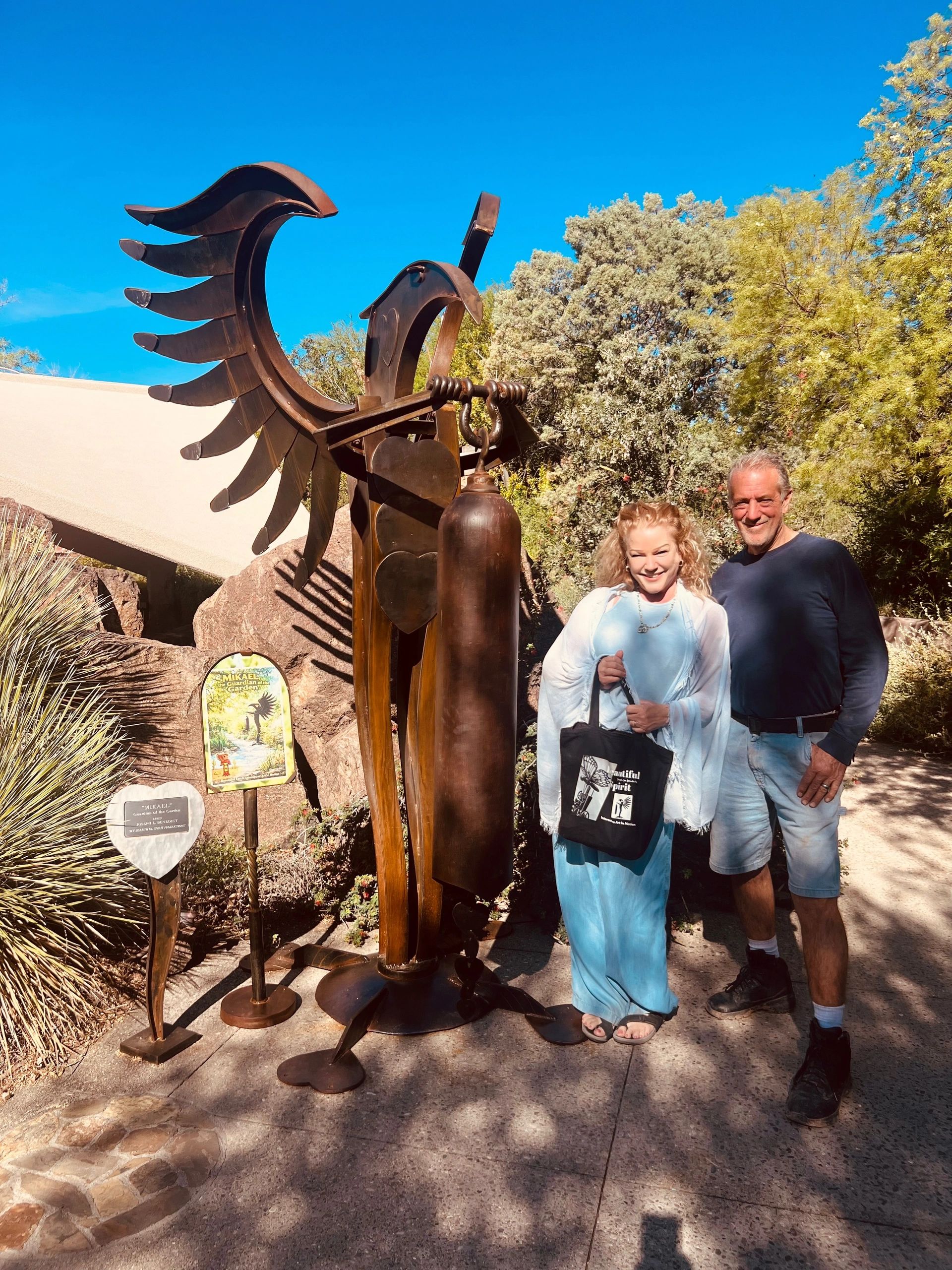 Two people posing beside a large metal angel sculpture outdoors on a sunny day.