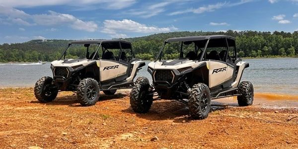Two beige Polaris RZR off-road vehicles parked by a lakeside under a blue sky.