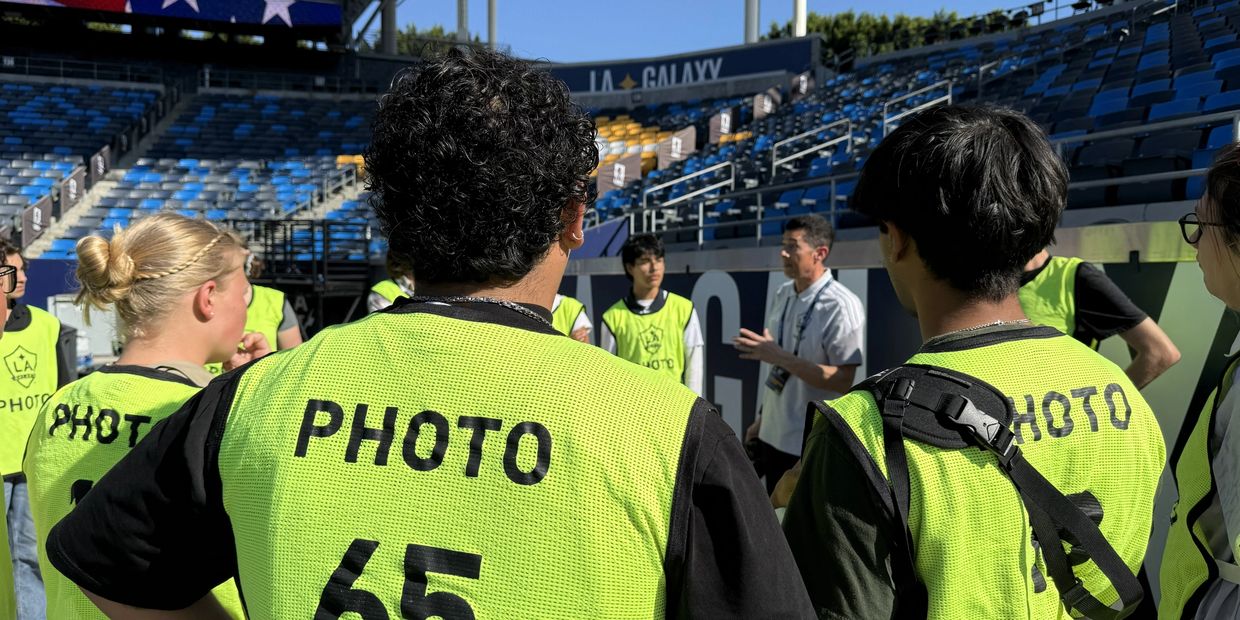 Group of photographers in yellow vests listening to instructions at a sports stadium.