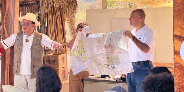 Two men present a map to an audience in an outdoor rustic setting.